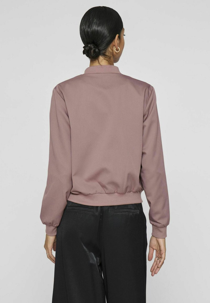 Rosa Bluse mit rundem Kragen, langen Ärmeln und elastischem Saum. Glatte Textur, minimalistisches Design, kombiniert mit schwarzen weit geschnittenen Hosen.