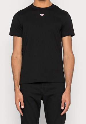 Mężczyzna ubrany w prostą czarną koszulkę z krótkim rękawem z małym czerwono-białym logo na środku klatki piersiowej, w połączeniu z czarnymi spodniami.