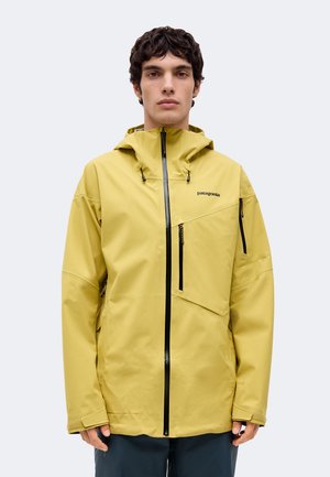 Un homme porte une veste jaune à capuche Patagonia, dotée d’une fermeture éclair sur toute la longueur et de plusieurs poches, sur un fond uni.