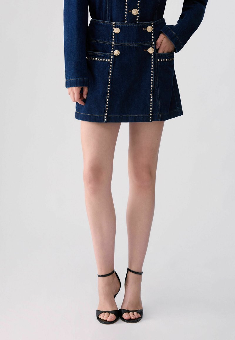 Femme portant une jupe en denim bleu foncé avec des boutons dorés et des clous, associée à des talons noirs à bout ouvert avec bride à la cheville, debout avec une main dans la poche.