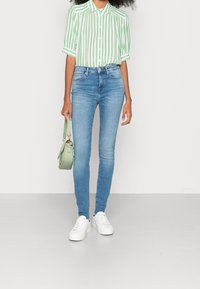 Mujer vistiendo una camisa de rayas verticales en verde y blanco, jeans ajustados de color azul claro, zapatillas blancas y sosteniendo un pequeño bolso de mano en color verde menta.