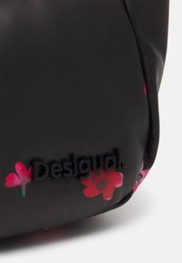 Gros plan d'un coin de sac noir texturé avec un imprimé floral rose et le logo "Desigual" noir en relief sur le tissu.