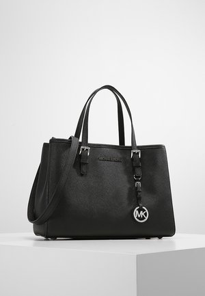 Sac cabas noir Michael Kors avec doubles poignées, bandoulière amovible et breloque logo MK sur un piédestal blanc.