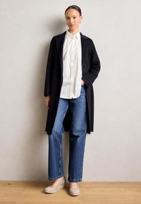 Navy wool coat, white button-up shirt, blue straight-leg jeans, and light gray slip-on shoes. Room with wooden floor and neutral wall.
