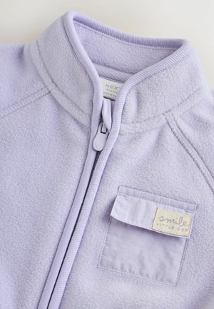 Jachetă din fleece lavandă cu guler înalt, închidere cu fermoar și buzunar la piept cu o etichetă pe care scrie „zâmbește micuțule.” Textură moale.