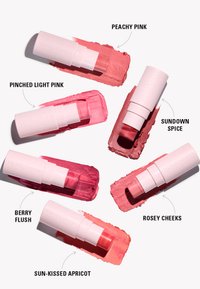 Seks leppestift-tuber i nyanser av rosa og fersken, arrangert med tilsvarende striper av kremet tekstur. Tubene har et hvitt design med ordet "REVOLUTION."