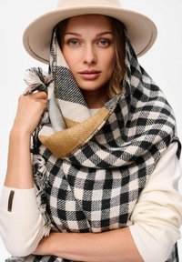 Czarna i biała szachownica szalik z kontrastującym złotym akcentem, pleciona tekstura, frędzle na krawędziach, noszony z beżowym kapeluszem i kremowym swetrem.