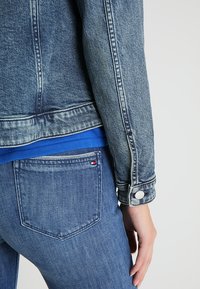 Giacca di jeans di colore blu sbiadito, con polsini abbottonati e dettagli sulle tasche, abbinata a una camicia blu visibile in vita e jeans di denim.