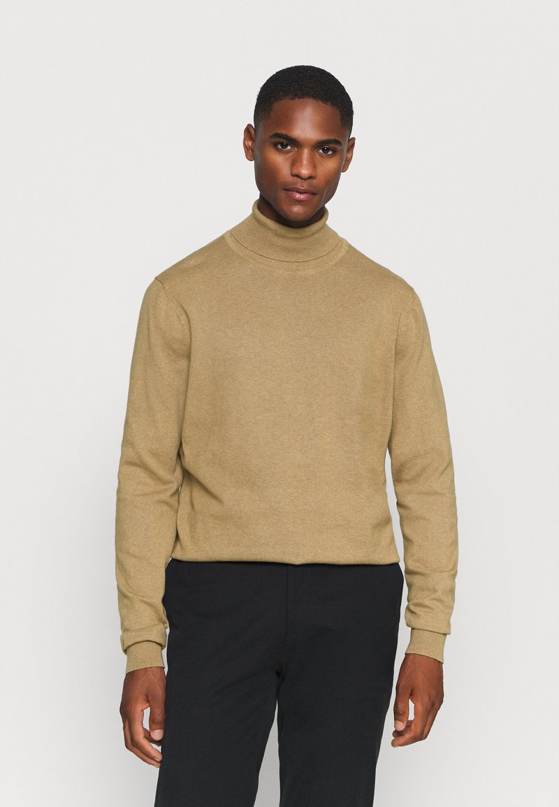 Beige turtleneck sweater lavet af blødt stof, med lange ærmer og en tætsiddende pasform. Sammen med sorte bukser.