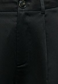 Gros plan sur un tissu noir montrant un bouton et une poche sur un pantalon avec des détails de couture visibles.