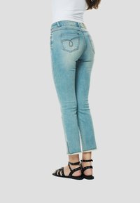 Világoskék farmernadrág, testhezálló fazonú, enyhén bokáig érő, gombos részletekkel és hátsó zsebekkel. Fekete szandállal viselve.