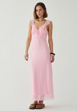 Frau trägt ein ärmelloses, bodenlanges pinkfarbenes Kleid mit Rüschendetails am Ausschnitt und beigen Plateau-Sandalen, steht vor einem schlichten Hintergrund.