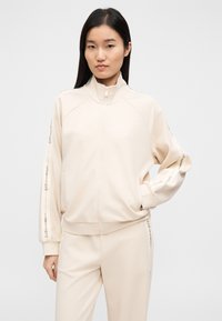 Svetlo bež zadrgovalna jakna z visokim ovratnikom, rebrastimi manšetami, sproščenim krojem in logotipom na straneh rokavov. Mehka tkanina, minimalen dizajn.