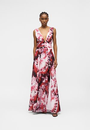 Γυναίκα που φοράει αμάνικο φόρεμα με βαθύ V λαιμόκοψη μέχρι το έδαφος, με σχέδιο βαφής κόκκινου, άσπρου και μαύρου, στέκεται μπροστά σε απλό λευκό φόντο.