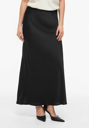 Femme portant une longue jupe noire en satin, un haut blanc et des talons noirs à bouts pointus, debout devant un fond clair.