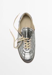 Silver metallisk sneaker med kviltad textur, krämfärgade snören, rund tå och en mjuk innervaddering. Platt sula med bruna accenter.