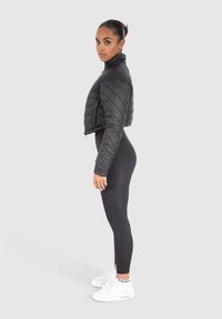 Zwarte cropped pufferjack met een geborduurd ontwerp, gecombineerd met zwarte leggings en witte sneakers. Glad textuur, aansluitende snit, ritssluiting.