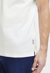 Balts polo krekls ar teksturētu audumu, sānu šķeltnēm un mazu trikoloru logotipa akcentu apakšmalā. Apvienots ar tumši zilām džinsām.