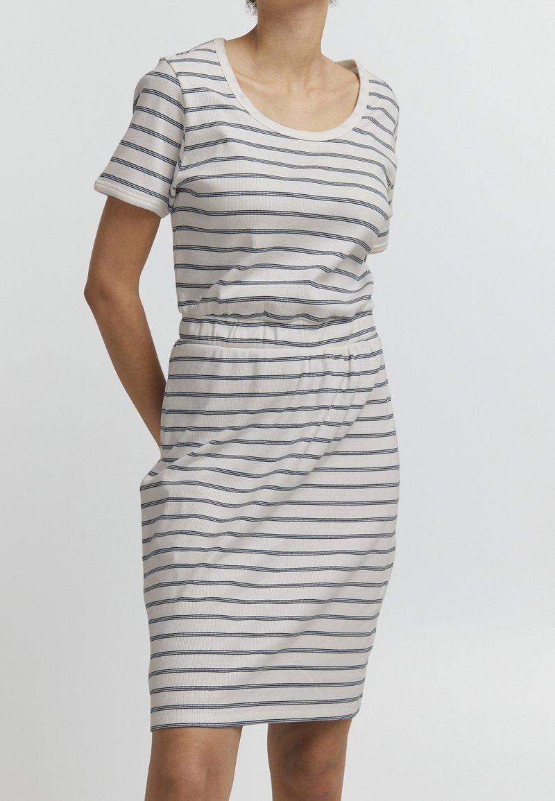 Femme portant une robe blanche à manches courtes, longueur genou, avec de fines rayures horizontales noires et une taille élastique, debout devant un fond uni.