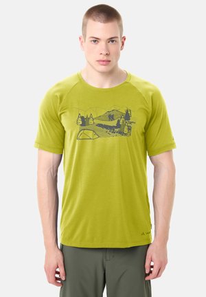 Uomo con t-shirt verde lime con grafica di montagna, alberi, escursionisti e tenda, abbinata a pantaloni verde oliva, in piedi su sfondo bianco.