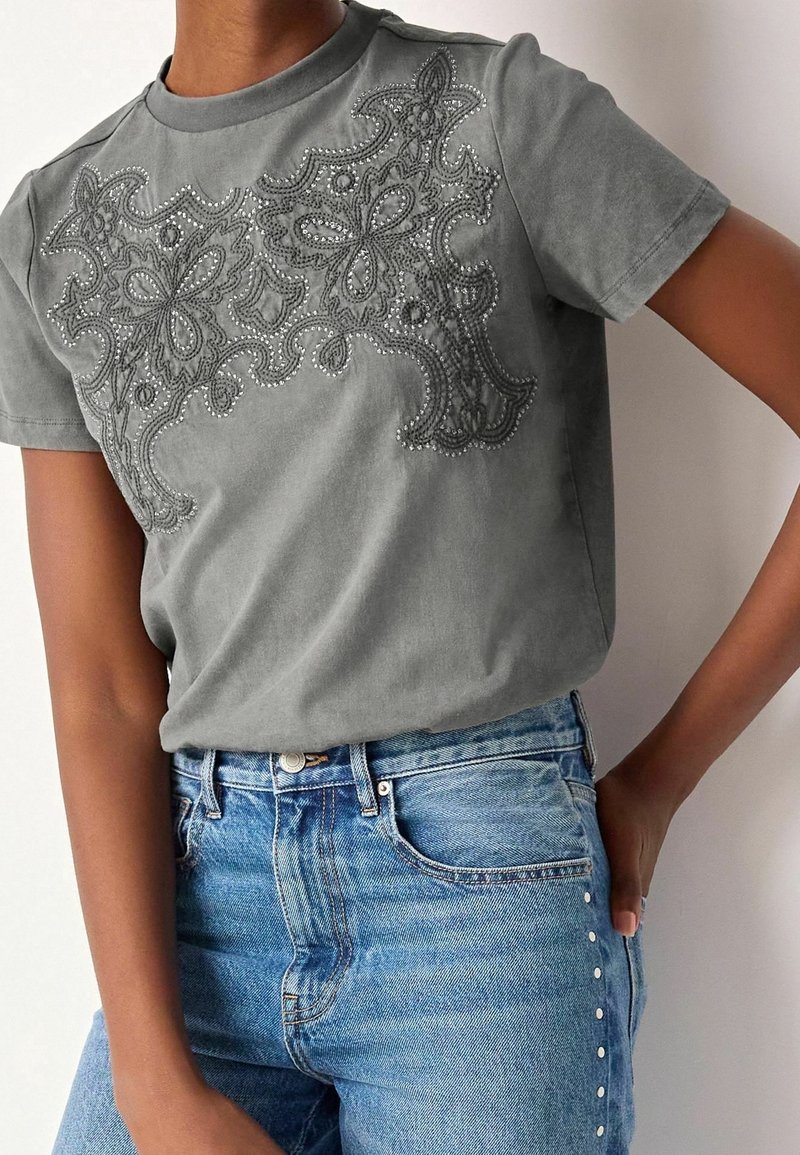 T-shirt gris à manches courtes avec une broderie élaborée et des perles sur le devant, associé à un pantalon en denim bleu avec des accents cloutés.