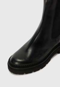 Bottine en cuir noir avec une finition lisse, bout rond, panneaux latéraux élastiques et semelle en caoutchouc texturée. Présente des surpiqûres décoratives le long du bord.