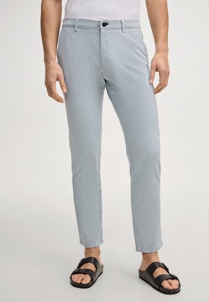 Mężczyzna ubrany w jasnoszare, obcisłe spodnie i czarne sandały na klamrę, stojący na jednolitym tle.