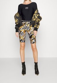 Svart kort sweatshirt, bomberjacka med guldaccent, mönstrade cykelshorts med guld- och vitdesign, samt svarta ankelstövlar med guldspetsar.