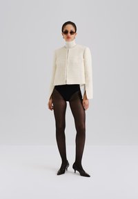 Texturerad cream croppad jacka med hög hals, dragen över en svart bodysuit och genomskinliga svarta strumpbyxor. Modellen bär mörka solglasögon och högklackat.