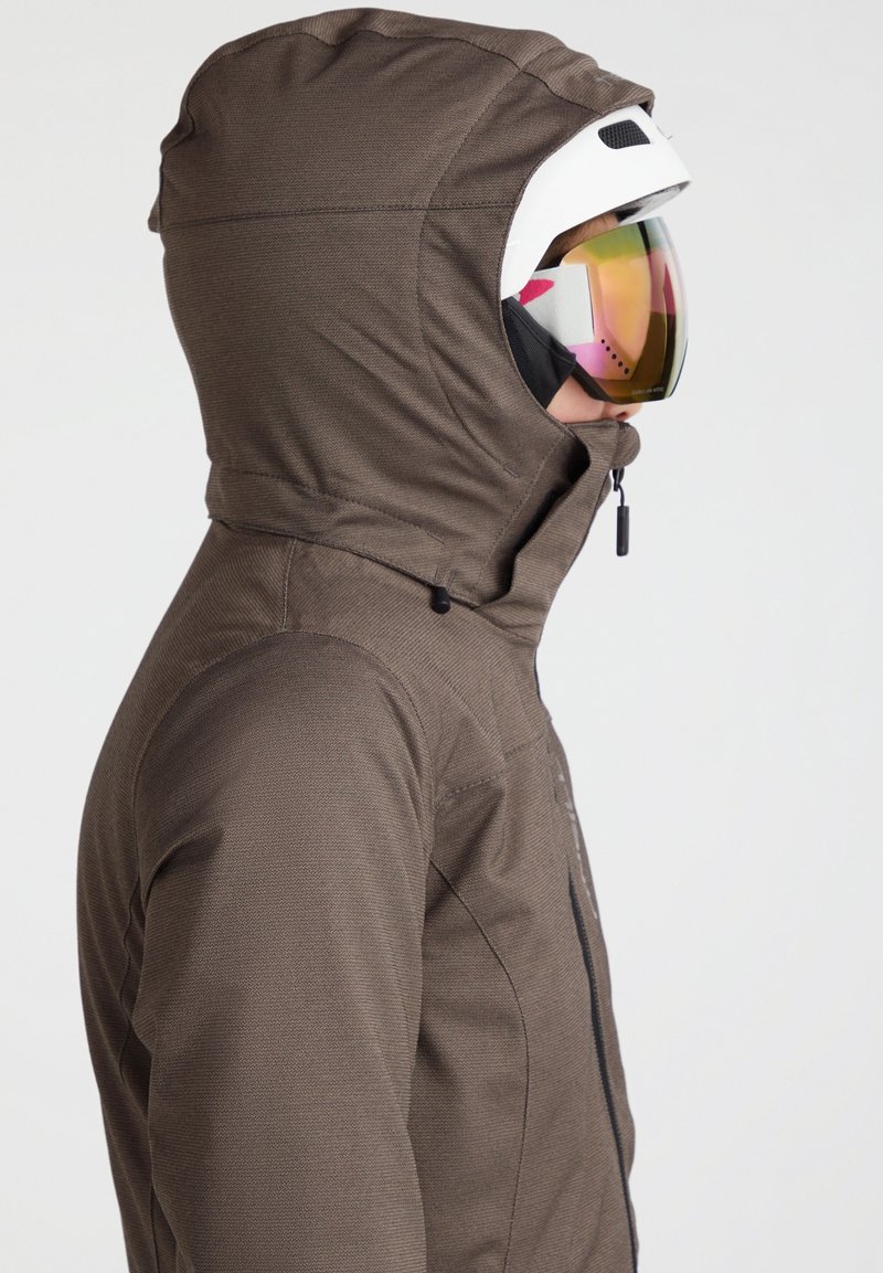 Skijacke aus braunem Stoff mit strukturiertem Finish, mit Kapuze, Reißverschluss und einer Person, die einen weißen Helm und farbenfrohe Skibrille trägt.