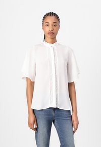 Blusa bianca con maniche corte a sbuffo, colletto arricciato e delicata rifinitura in pizzo lungo la parte anteriore, abbinata a jeans blu.