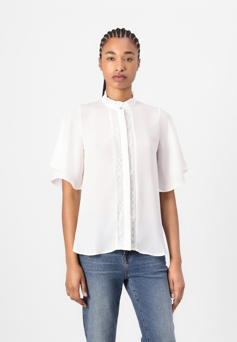 Blusa bianca con maniche corte a sbuffo, colletto arricciato e delicata rifinitura in pizzo lungo la parte anteriore, abbinata a jeans blu.