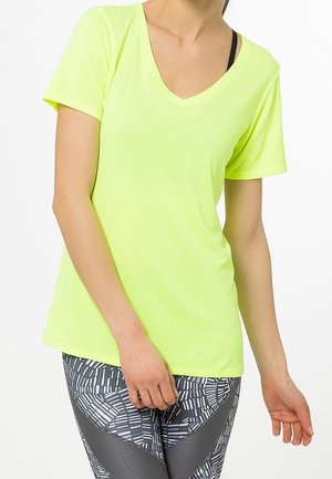 Kvinde iført en neon gul T-shirt med korte ærmer og V-hals samt mønstrede sorte og hvide leggings, stående med den ene hånd hvilende ved hoften.
