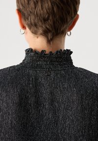 Tessuto nero testurizzato con strisce metalliche verticali, caratterizzato da un colletto arruffato e un dettaglio con bottone sul retro.