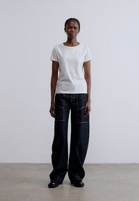 Fehér pamut póló laza, sötét farmernadrággal, amely elején zsebekkel és kontrasztos varrással rendelkezik, fekete cipővel viselve.