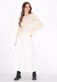 Crème gebreide trui met een opengewerkt patroon, gecombineerd met een vloeiende witte maxi-jurk en zwarte enkellaarsjes met gespaccenten.