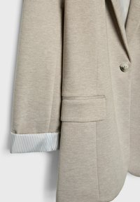 Manche de blazer beige texturé montrant un seul bouton, une poche à rabat, et un poignet de chemise rayée dépassant de l'extrémité de la manche.