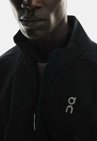 Black jacket with a high collar, featuring a silver logo on the chest. The fabric appears smooth and slightly shiny. Zippered front closure.
