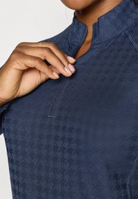 Marineblå zip-up genser med høy krage, med et teksturert houndstooth-mønster. Stoffet ser jevnt ut og er noe elastisk.