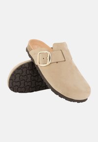 Beige mokkasiner i semsket skinn med en bred stropp og gullspenne, med en teksturert svart gummisåle og en glatt indre fôr.