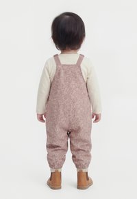 Bambino in piedi di spalle che indossa una camicia crema a maniche lunghe, una salopette rosa con motivo floreale e stivaletti color beige su sfondo bianco.
