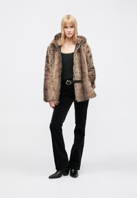 Brun jakke i imiteret pels med hætte, lynlås og sidelommer; sort tætsiddende top, sorte fløjlshosen og ankelstøvler.