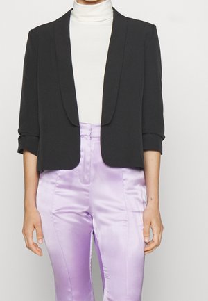 Person wearing a white turtleneck, cropped black blazer with rolled sleeves, and high-waisted shiny lavender pants.