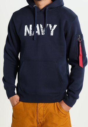 Mężczyzna w granatowej bluzie z kapturem z białym napisem "NAVY" i kieszenią na zamek z czerwoną metką na lewym rękawie, w połączeniu z musztardowymi spodniami.