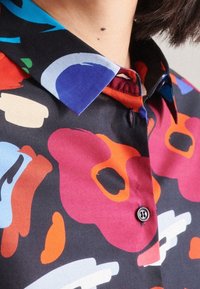 Chemise multicolore avec une base noire, présentant des formes abstraites en rouge, bleu et orange. Détails remarquables comprenant un col pointu et un bouton noir.