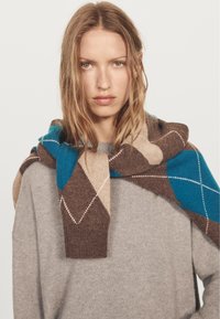 Bruine, beige en turkooizen sjaal met een patroon van ruitvormen en witte stiksels, over een grijze trui gedrapeerd. Zachte textuur, gemiddelde dikte.