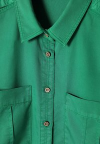 Cămașă verde, cu nasturi, din bumbac, având un guler clasic, două buzunare frontale și nasturi maro, cu o textură netedă.