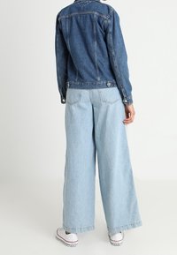 Jeansjacke mit tailliertem Schnitt, kombiniert mit weit geschnittenen, hellblauen Jeans. Das Outfit hat einen klassischen Kragen und Knopfdetails. Weiße Sneaker runden den Look ab.