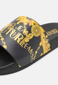 Sandale slide noire avec des motifs floraux et chaînes dorés ornés ainsi qu’un texte doré affichant "Versace Jeans Couture" sur la sangle.