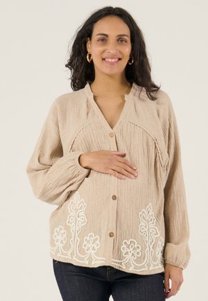 Lächelnde schwangere Frau in beige strukturierter Bluse mit weißer Blumenstickerei, die ihre Hand auf den Bauch legt vor einfachem Hintergrund.